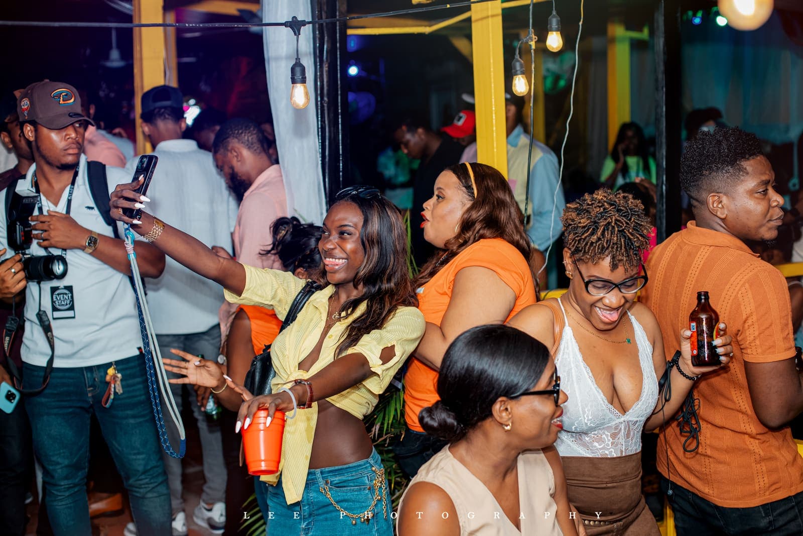 The floor at UNCUT Vol. 1 in Pétion-Ville: guests laughing under filament bulbs, a phone up, drinks in hand.